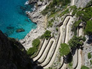 Cette photo est prise par drone sur l'île de Capri, près de Naples, en Italie. On y voit un chemin en pierre beige serpentant jusqu'à une petite plage rocheuse aux eaux turquoises et limpides